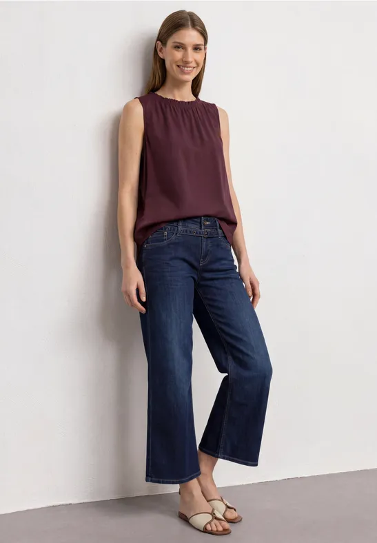 Frau in lila Top, weiten blauen Jeans und Sandalen steht an der Wand und lächelt.