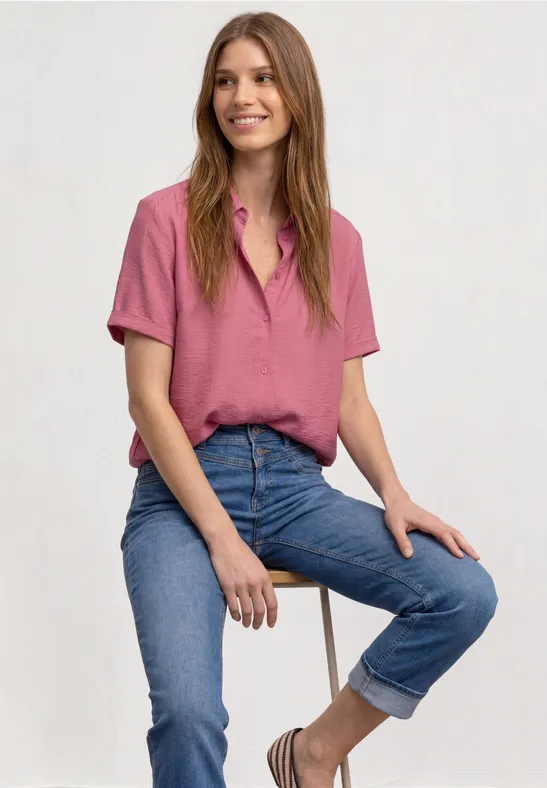 Een glimlachende vrouw zit op een kruk, draagt een roze blouse en opgerolde blauwe spijkerbroek.