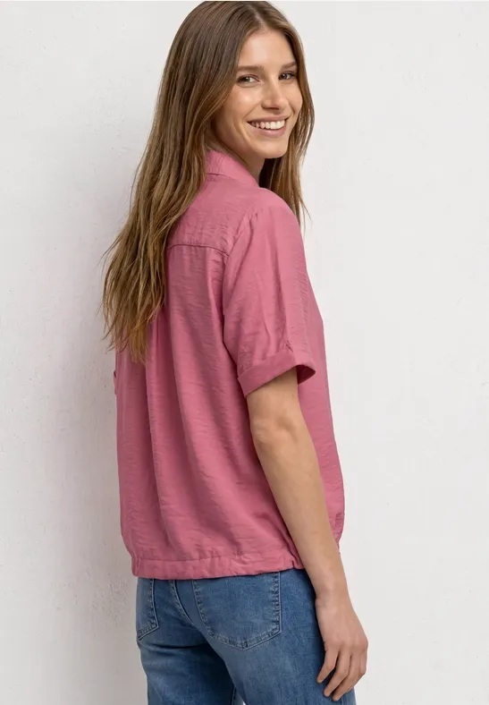 Een vrouw in roze kort mouwige blouse en spijkerbroek kijkt glimlachend over haar schouder tegen een witte achtergrond.