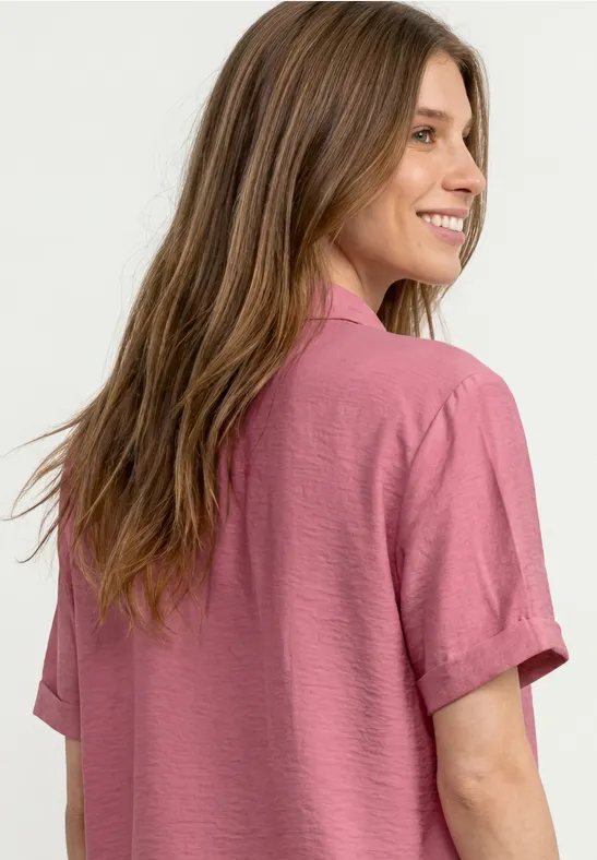 Een vrouw met lang bruin haar draagt een roze overhemd en lacht terwijl ze zich omdraait.