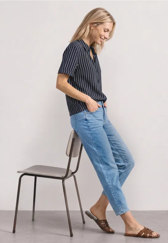Vrouw in een gestreept overhemd, blauwe jeans en sandalen leunt nonchalant tegen een stoel.