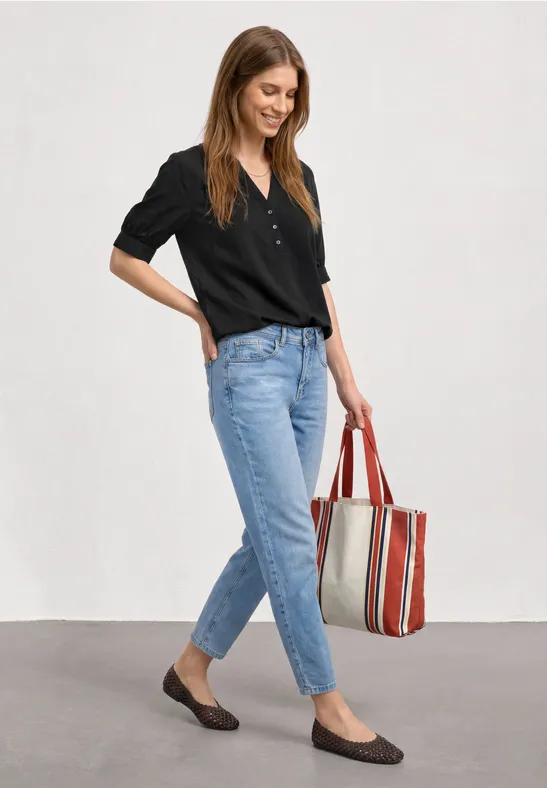 Frau in schwarzer Bluse, Jeans und flachen Schuhen hält gestreifte Tasche.