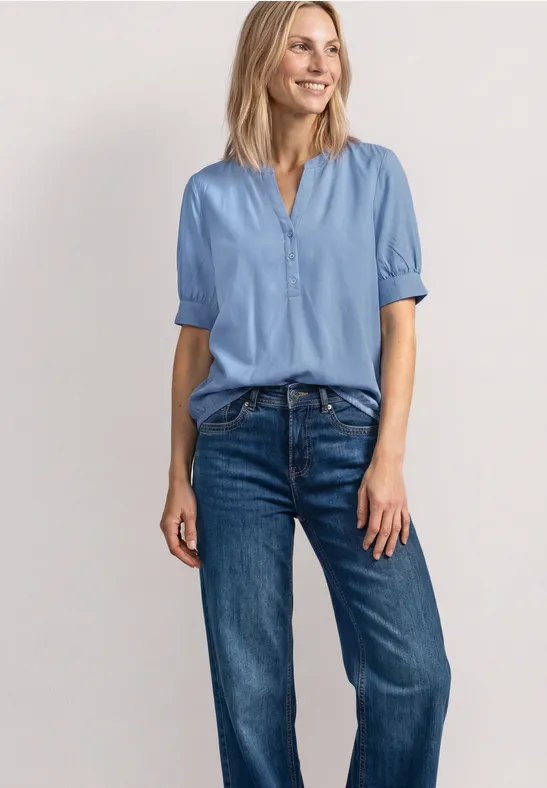 Frau in hellblauer Bluse und blauen Jeans vor hellem Hintergrund.