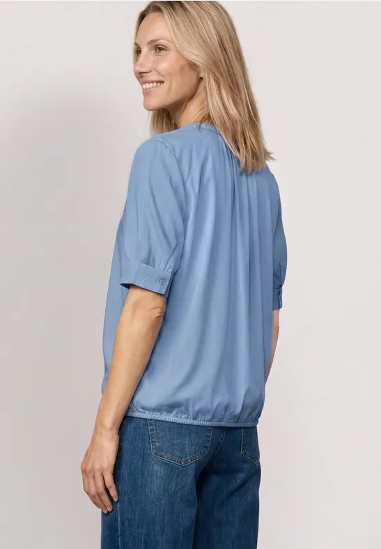 Frau steht von hinten, trägt hellblaue Bluse und Jeans.