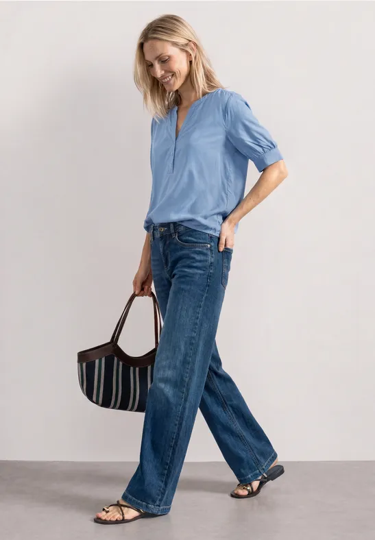 Frau in hellblauer Bluse, weiten Jeans, Sandalen und gestreifter Handtasche vor neutralem Hintergrund.