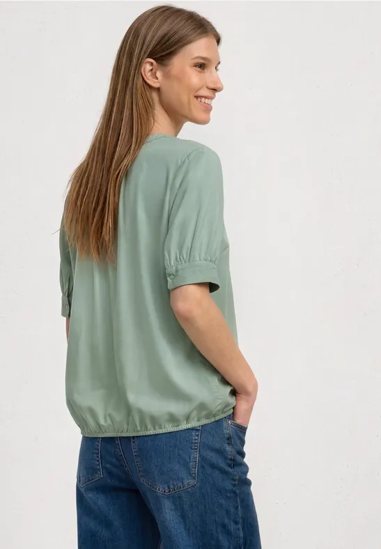 Frau mit langen Haaren von hinten, trägt grünes Oberteil und blaue Jeans, Hände in den Taschen.