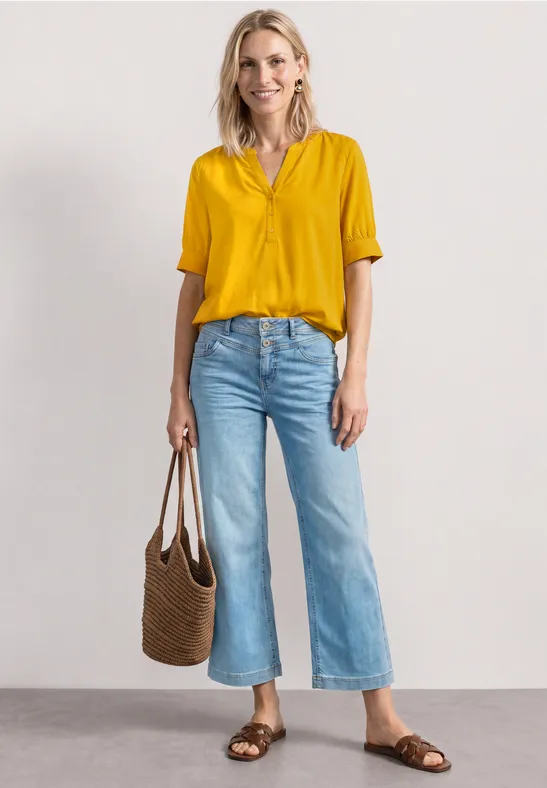 Frau in gelber Bluse, weiten Jeans, braunen Sandalen und geflochtener Tasche.