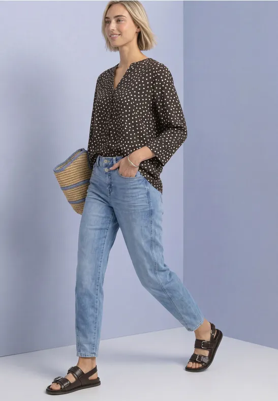 Eine Frau in Jeans, schwarzer gepunkteter Bluse und Sandalen trägt eine Strohtasche vor lila Wand.