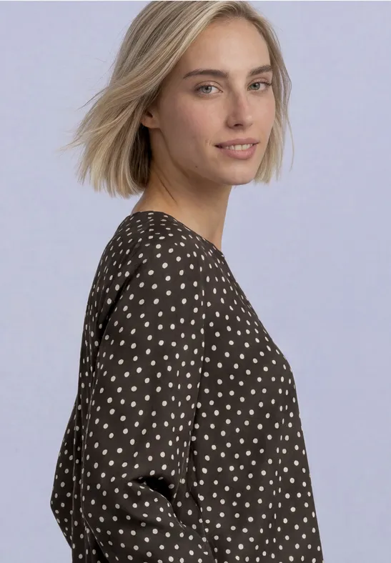Eine blonde Frau schaut über die Schulter, trägt ein schwarzes Kleid mit weißen Punkten.