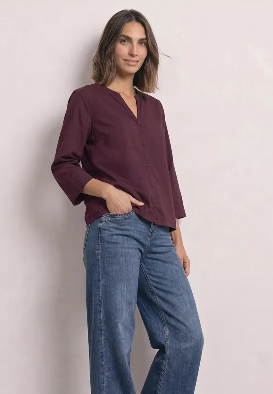 Frau in weinroter Bluse und weitem Jeans, lässig stehend vor hellem Hintergrund.
