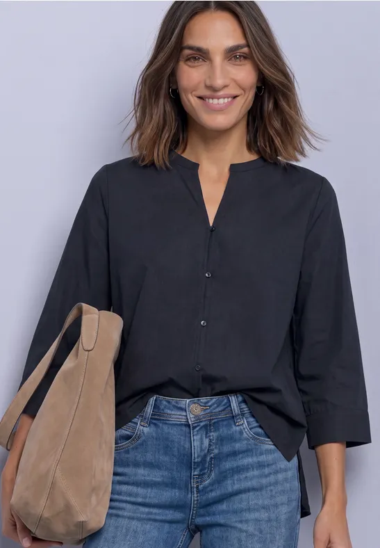 Frau mit schwarzer Bluse, blauen Jeans und beigem Schulterbeutel lächelt vor hellgrauem Hintergrund.