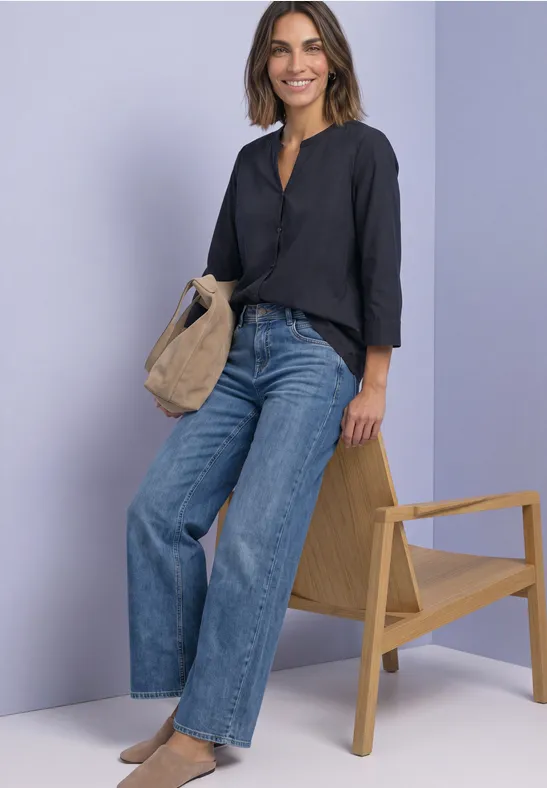 Frau in schwarzer Bluse und weitem Jeans, mit beiger Tasche neben einem Sessel vor lila Hintergrund.