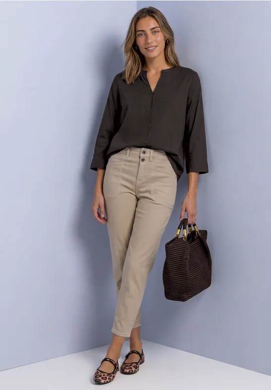 Eine Frau in schwarzer Bluse, beigen Hosen und Sandalen hält eine braune Tasche lächelnd.