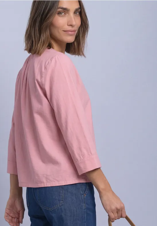Frau trägt locker sitzende rosa Bluse mit 3/4-Ärmeln und blaue Jeans, Blick über die Schulter.