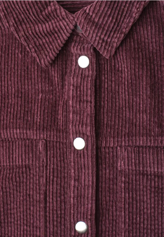 Nahaufnahme einer weinroten Cordjacke mit Struktur, Hemdkrause und weißen Druckknöpfen.
