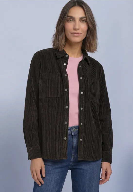 Kobieta na neutralnym tle w brązowym overshirt z sztruksu, różowym topie i jeansach.
