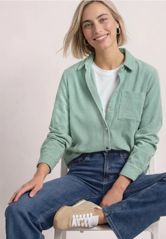 Frau sitzt im Schneidersitz, lächelt, trägt mintgrünes Hemd, weißes T-Shirt, Jeans und beige Sneaker.