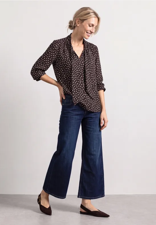 Frau in gepunkteter Bluse, weiten cropped Jeans und flachen Schuhen.
