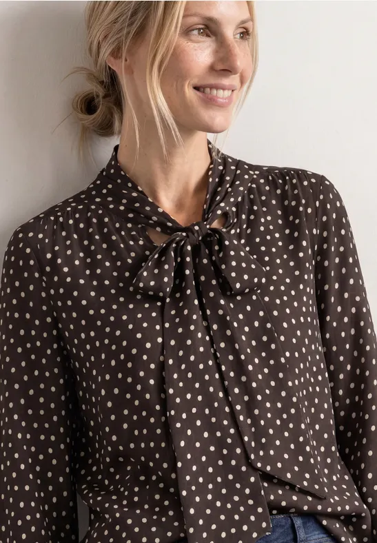 Blonde Frau in brauner Bluse mit weißem Herzmuster und Schleife am Kragen, lächelnd zur Seite blickend.