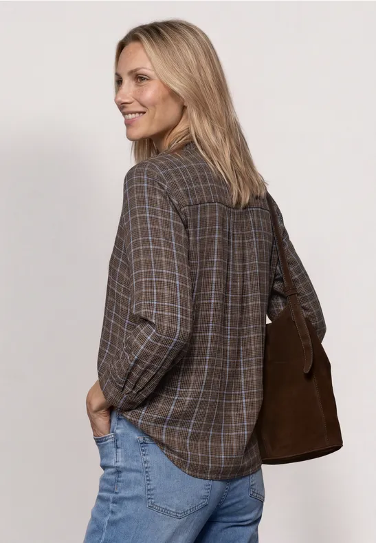 Frau mit blonder Mähne trägt kariertes Hemd, Jeans und eine braune Schultertasche, lächelt seitlich.
