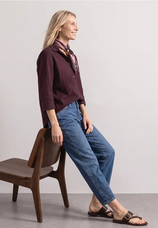 Eine Frau lehnt an einem Stuhl, trägt Jeans, bordeauxfarbenes Hemd, schwarze Sandalen und lächelt zur Seite.
