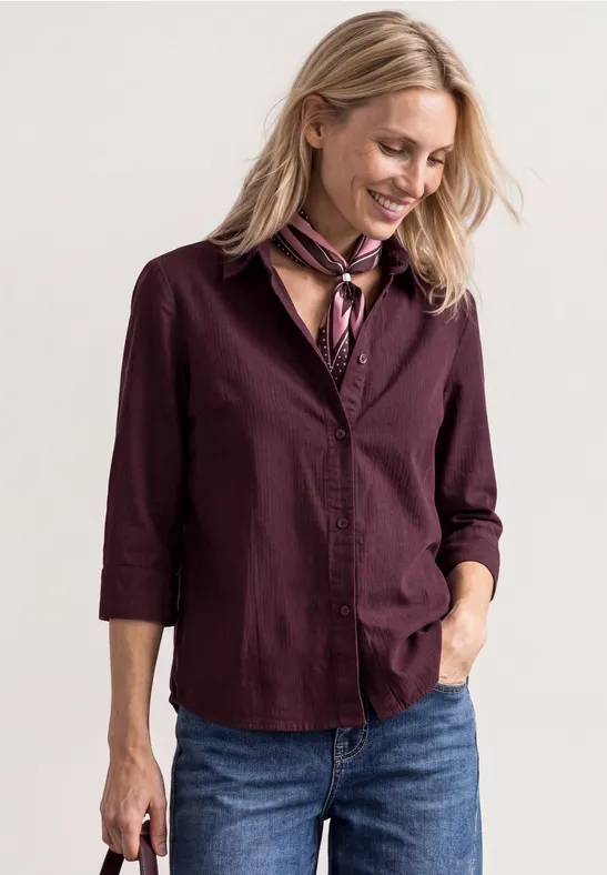 Frau in bordeauxfarbener Bluse, Jeans und Halstuch, lächelnd und nach unten blickend