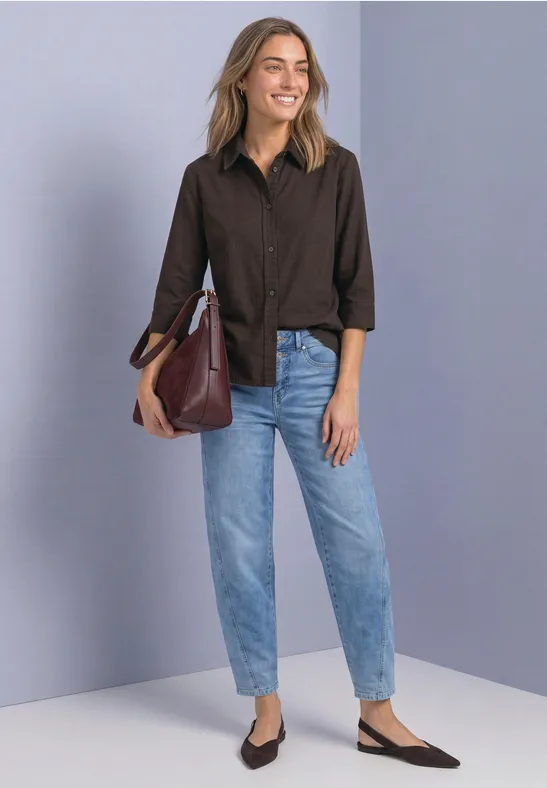 Eine Frau in braunem Hemd, hellblauen Jeans, schwarzen Slingback-Schuhen und großer bordeauxfarbener Tasche vor grauer Wand.