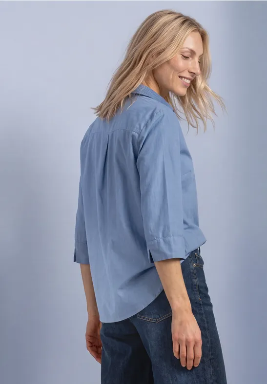 Frau in hellblauem Hemd und Jeans, lächelt seitlich zur Kamera.
