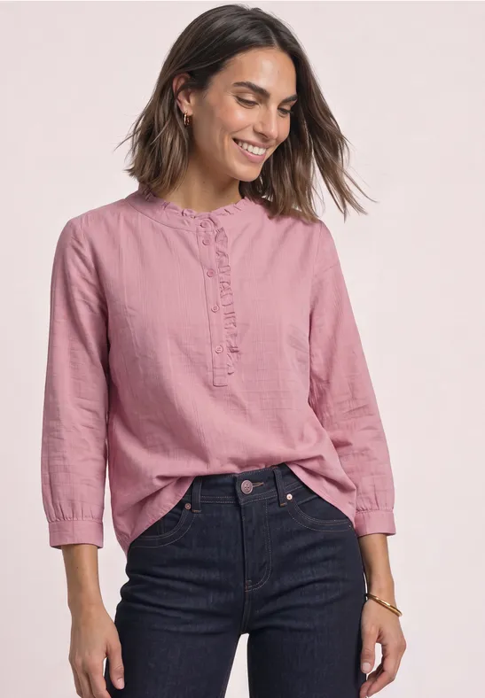 Frau in rosa Bluse und dunklen Jeans, lächelt vor hellem Hintergrund.