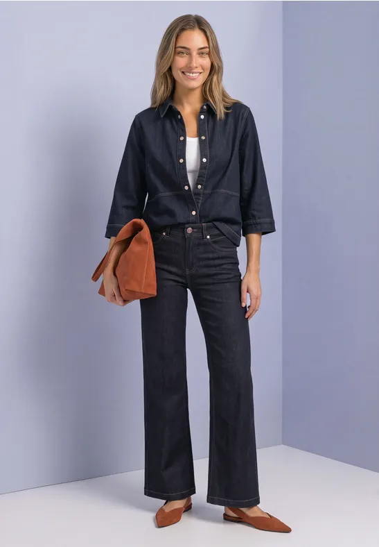 Frau in dunklem Denim-Hemd und Schlaghose mit weißem Top, brauner Tasche und spitzen Schuhen vor hellem Hintergrund.