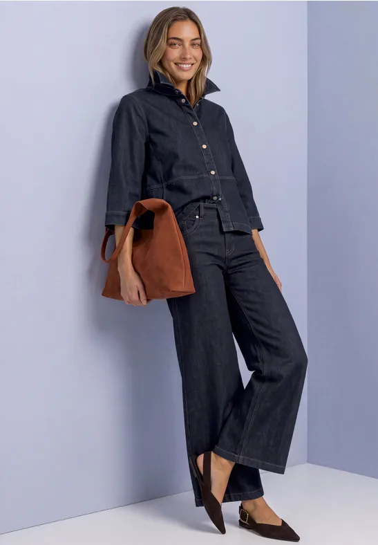 Frau in dunklem Denim-Outfit mit weiter Hose, Jeansjacke, schwarzen Schuhen und brauner Tasche vor hellem Hintergrund.