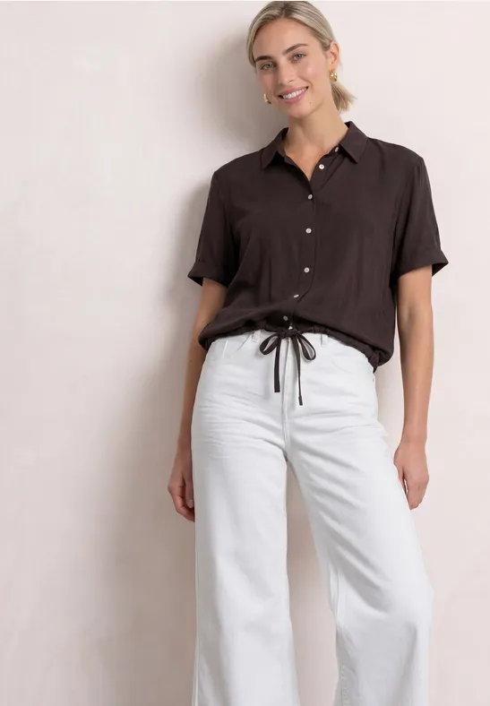 Vrouw in losse bruine blouse en wijde witte broek met taillekoord.