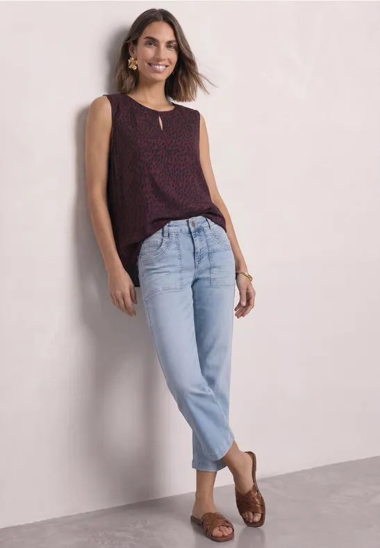 Junge Frau lehnt an einer Wand, trägt ärmelloses gepunktetes Top, helle Jeans und braune Sandalen.