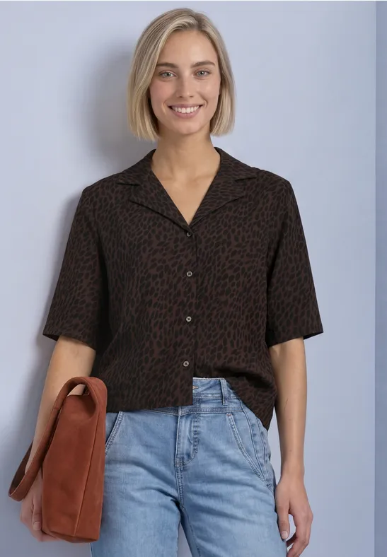Junge Frau in kurzärmeligem, braun-schwarzem Print-Hemd, Jeans und Umhängetasche, lächelnd