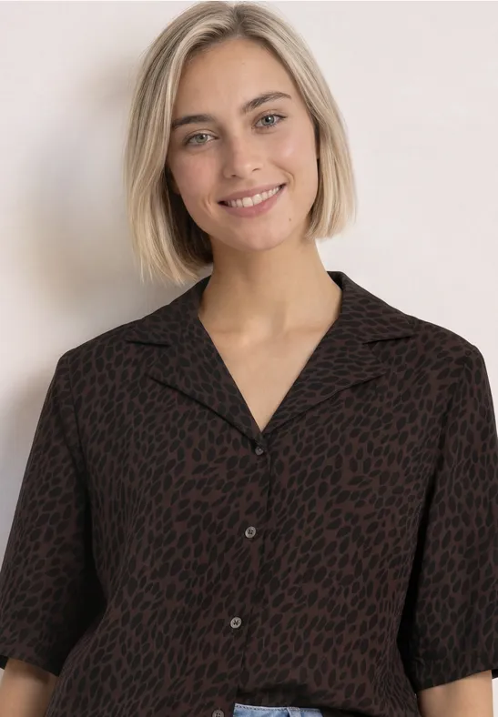 Junge Frau mit kurzem blonden Haar lächelt in einer dunkel gemusterten Bluse vor hellem Hintergrund.