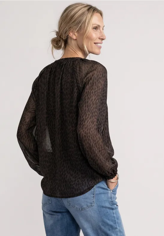 Frau in schwarzer, halbtransparenter Bluse mit dezentem Muster und Jeans, von hinten lächelnd.