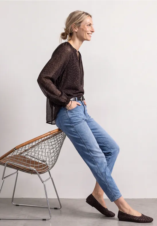 Frau in dunkler Bluse und Jeans sitzt lächelnd seitlich auf einem Drahtstuhl vor hellem Hintergrund.