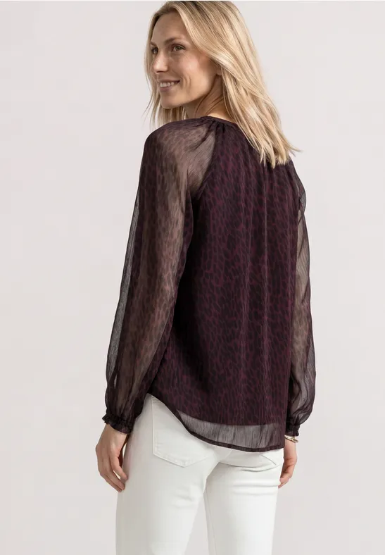 Eine Frau trägt eine dunkelrote Bluse mit transparenten schwarzen Ärmeln und weiße Hose.