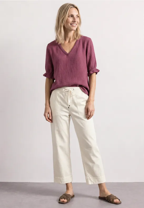 Eine lächelnde Frau in roter Bluse, weißer Culotte und Sandalen vor neutralem Hintergrund.