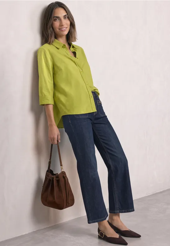 Frau in grüner Bluse und Jeans, braune Tasche, lässig an Wand gelehnt.
