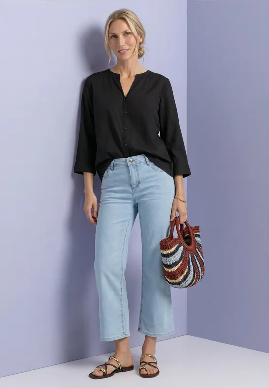 Frau mit schwarzer Bluse, hellblauen Jeans, gestreifter Tasche und Sandalen vor lila Hintergrund.