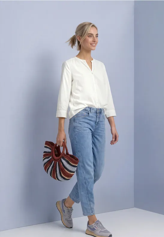 Junge Frau in weißer Bluse und Jeans posiert vor pastellfarbener Wand und hält eine bunte Tasche.