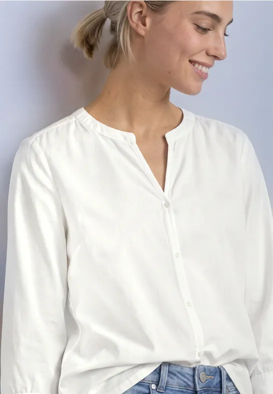 Eine lächelnde Frau trägt eine weiße Bluse mit Knopfleiste und Jeans.