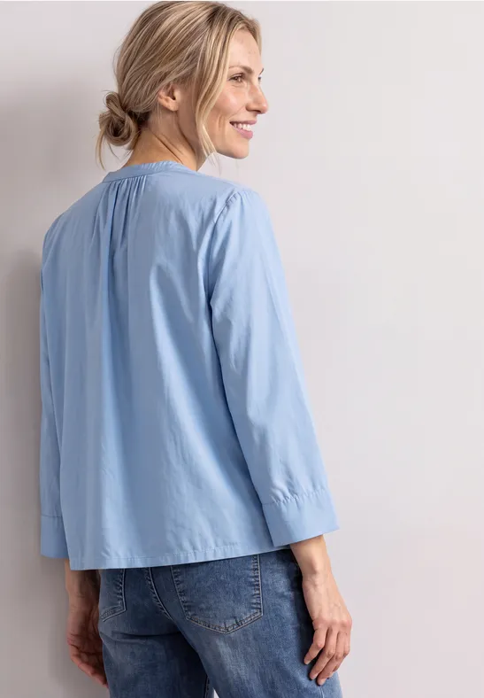 Lächelnde Frau in hellblauer Bluse und Jeans, seitlich stehend vor hellem Hintergrund.
