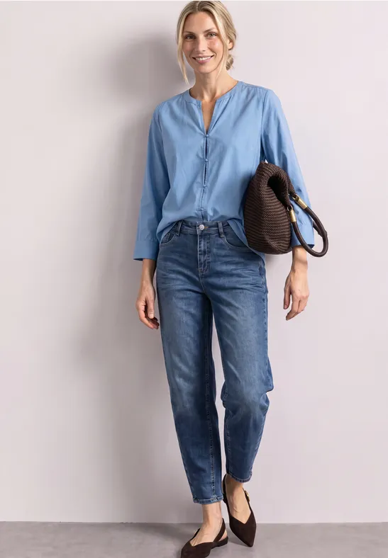 Junge Frau vor heller Wand, trägt blaue Bluse, Jeans und schwarze Ballerinas, hält eine Tasche.