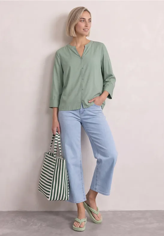 Frau mit kurzem blondem Haar, grünem Top, hellblauen Jeans, grünen Sandalen und gestreifter Tasche vor schlichter Kulisse.