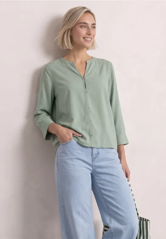 Frau mit blondem Bob in hellgrüner Bluse mit V-Ausschnitt und heller Jeans, lächelnd vor hellem Hintergrund.