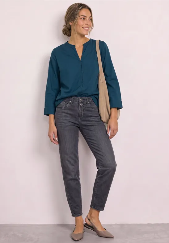 Frau trägt petrolfarbene Bluse, schwarze Jeans, flache Schuhe und beigetönige Schultertasche, sie lächelt stehend.