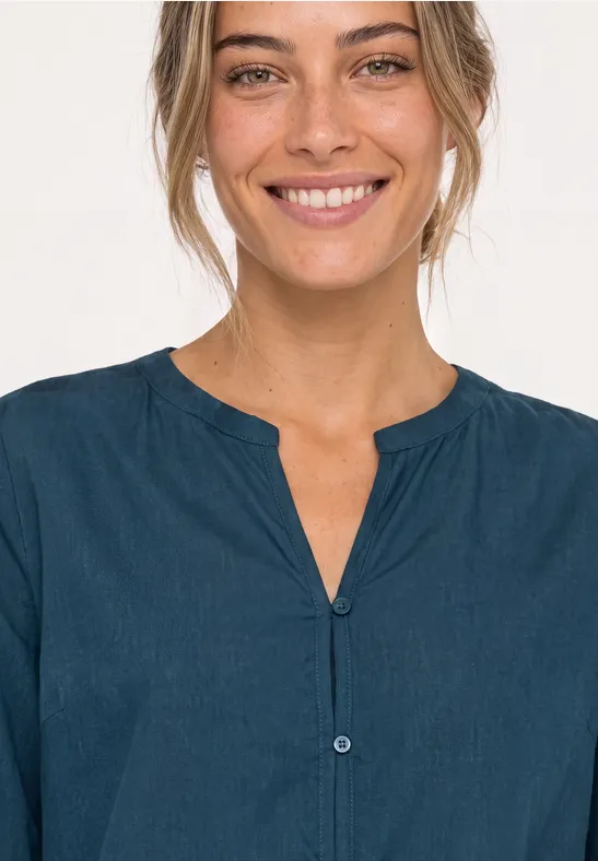 Eine lächelnde Frau mit dunkelblauer, kragenloser Bluse vor hellem Hintergrund.