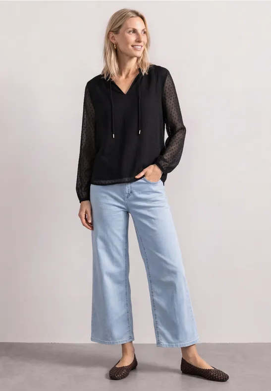 Kobieta w czarnej przezroczystej bluzce z kapturem, jasnych szerokich dżinsach i płaskich butach.